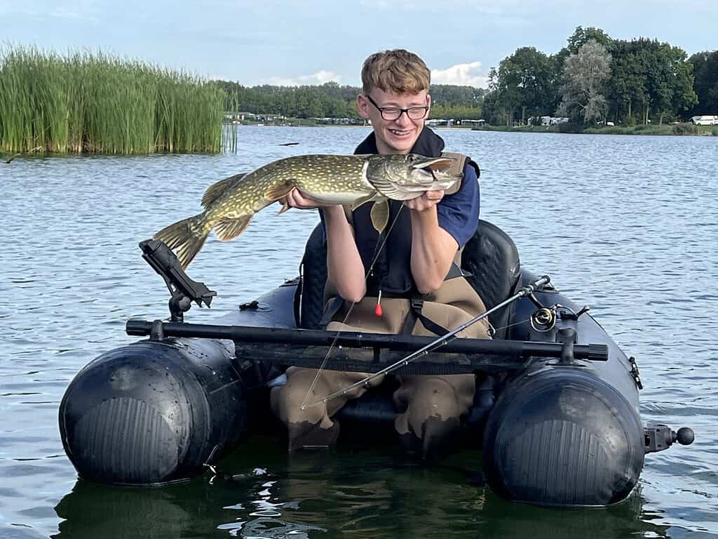 Een persoon zit in een kleine bellyboat en houdt een grote vis omhoog die hij heeft gevangen op de Jeugddag van Sportvisserij Zuidwest Nederland, met riet en bomen op de achtergrond.