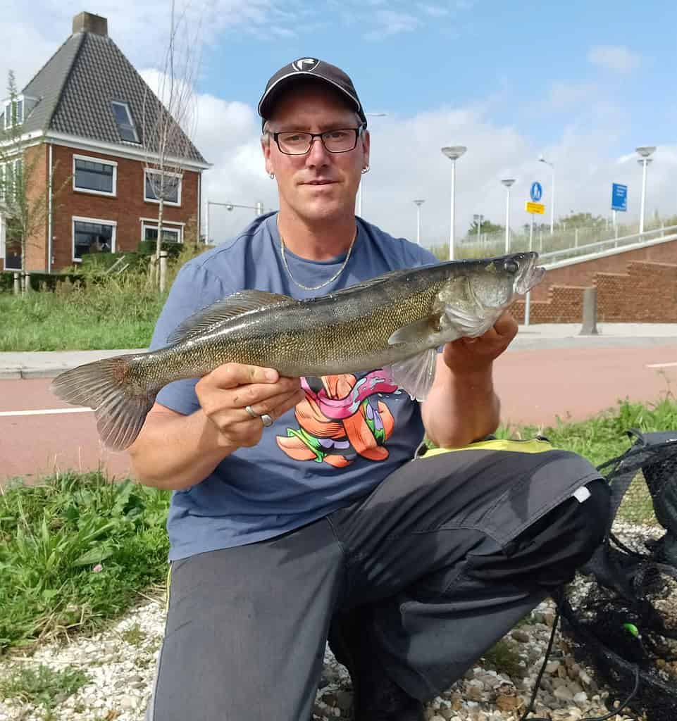 Een persoon met een pet en een bril zit buiten, trots een van de mooie vangsten vasthoudend. Er is een bakstenen huis en wat trappen op de achtergrond.