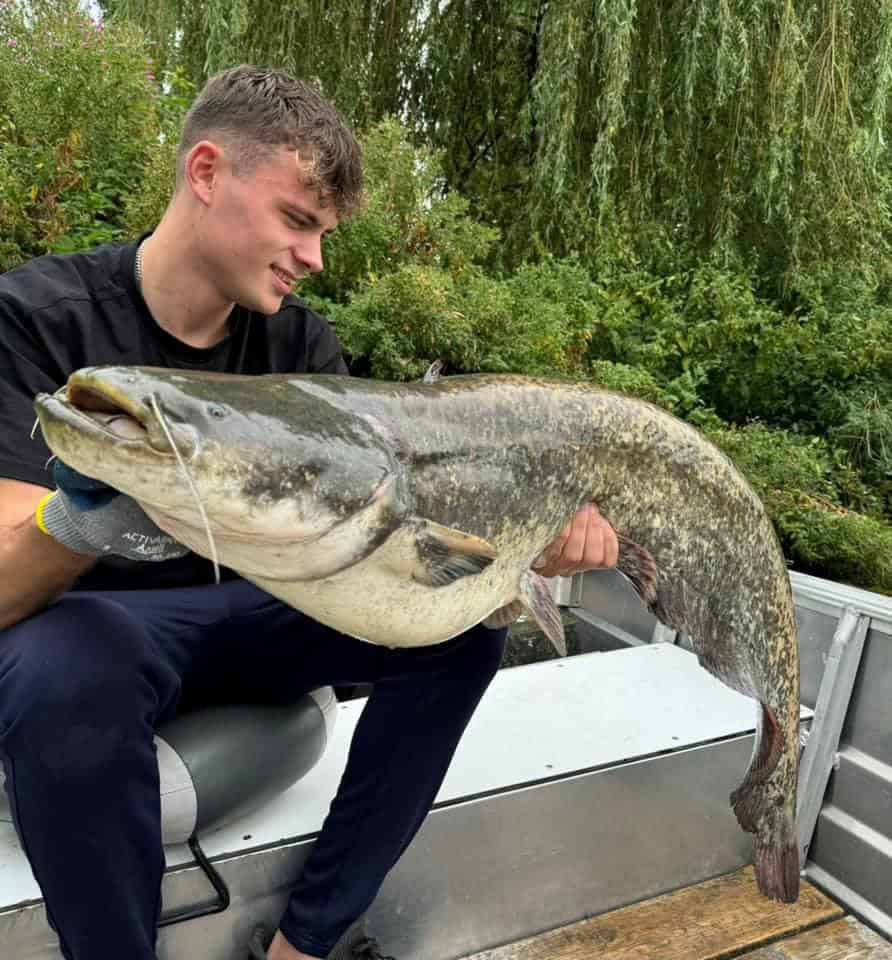 Een man zit in een metalen bootje en houdt met beide handen een grote vis vast. Op de achtergrond is een van de prachtige vangsten met groen te zien.