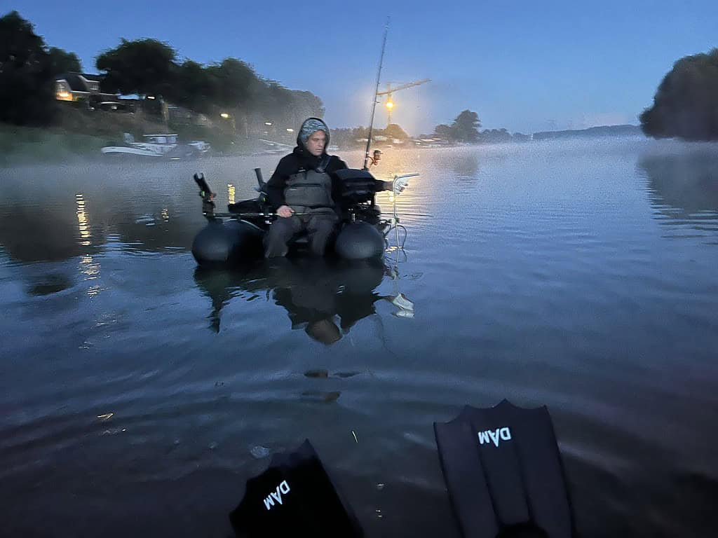 Een persoon gekleed in visgerei zit op een opblaasbare boei met een hengel op een mistig wateroppervlak bij zonsopgang, in de hoop een XXL-meerval te vangen, net zoals Paul Breems deed tijdens zijn visexpedities.