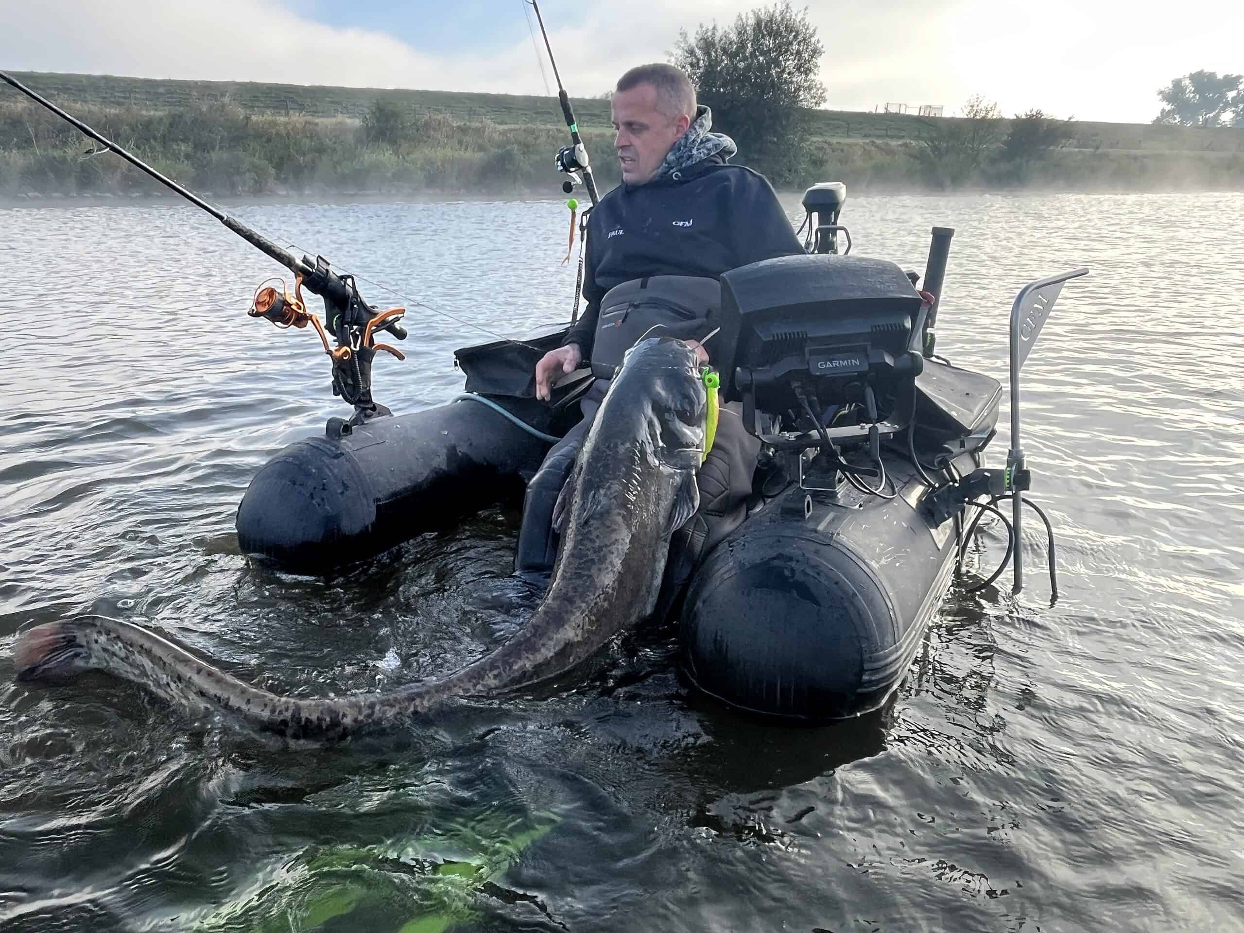 Een persoon in een zwarte opblaasbare boot houdt een grote vis vast in een mistig meer, met hengels die uit de boot steken. Op naar de twee meter lijken ze vastbesloten om hun geluk te beproeven voor een nog grotere vangst.