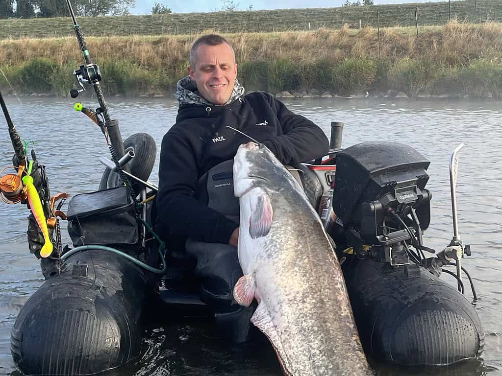 Een persoon zit in een opblaasbare boot op een rivier, lachend en met een vangsten-waardige grote meerval in zijn hand. Visgerei is zichtbaar in de boot en er is een grasachtige oever op de achtergrond.