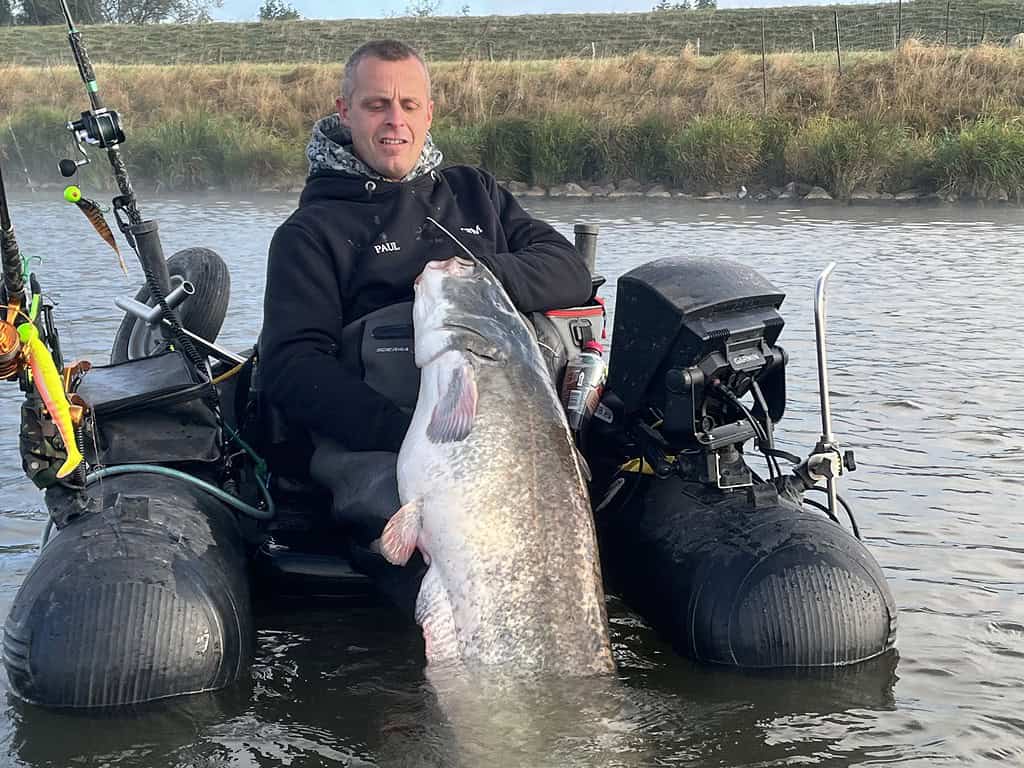 Paul Breems zit in een opblaasboot, trots een XXL-meerval vasthoudend. De boot drijft op kalm water met grasachtige oevers op de achtergrond—een serene scène van vissen op zijn best.