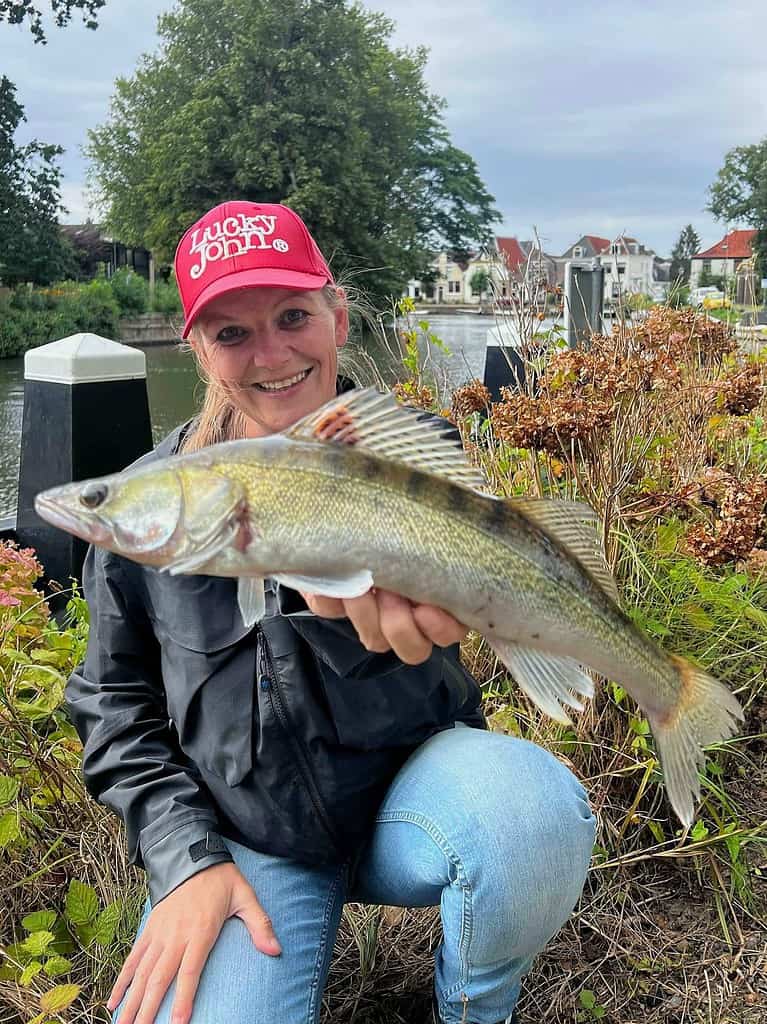 Persoon in een zwart jasje en een rode pet die een vis vasthoudt, poseert bij een waterpartij met huizen en groen op de achtergrond. Hij laat een van de schitterende vangsten van deze week zien.