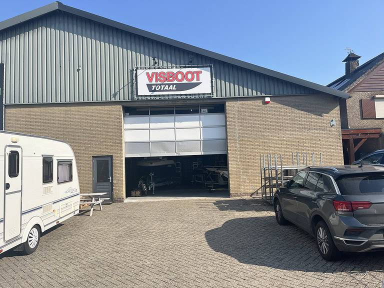 Een loods met een bordje "VISBOOT TOTAAL" boven een open garagedeur, waaruit boten blijken. Een caravan en een auto staan op de voorgrond van het nieuwe pand op een zonnige dag.