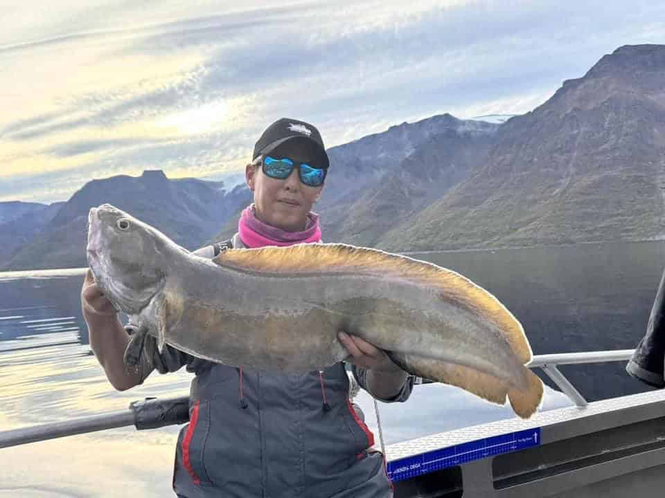 Persoon op een boot die een grote vis vasthoudt met een bergachtig landschap en een kalm meer op de achtergrond. Met een zonnebril, een pet en buitenkleding tonen ze trots hun vangsten van de week.