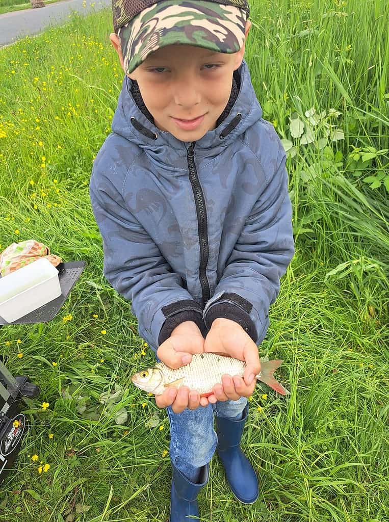 Een kind met een blauw jasje en een camouflagepet houdt een kleine vis met beide handen vast terwijl hij in een grasveld staat. Het toont een van de memorabele vangsten van de week.