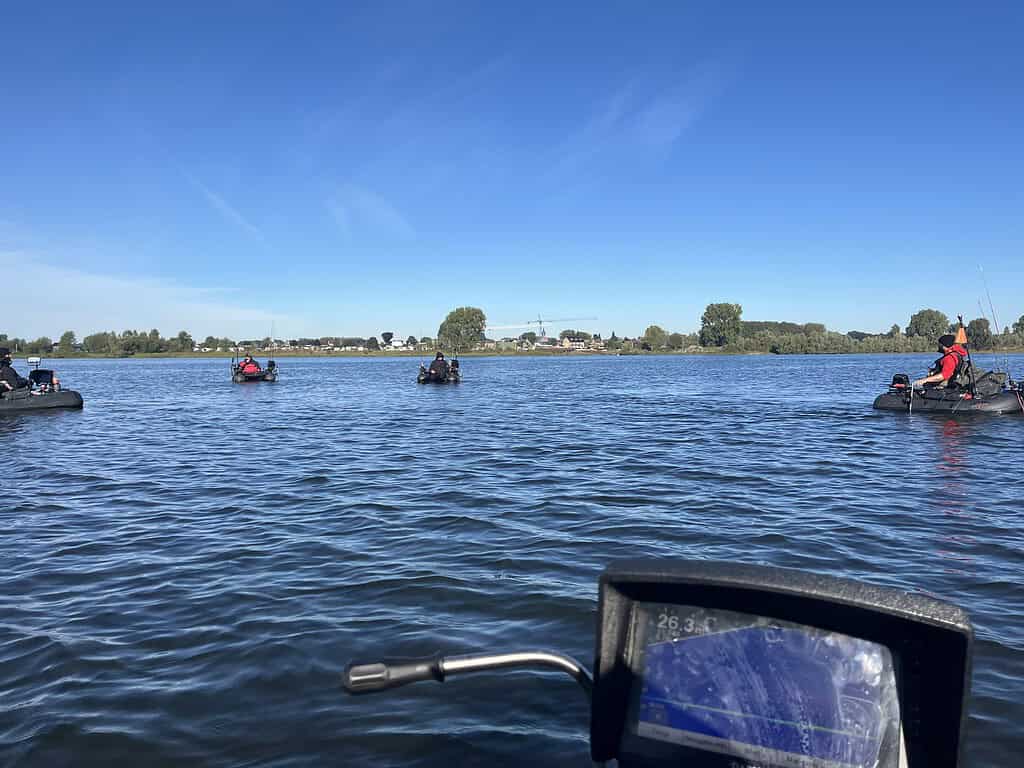 Meerdere mensen vissen op kleine boten in een kalm meer onder een helderblauwe lucht. Een fishfinder-scherm, dat dieptemetingen in twee meter weergeeft, is zichtbaar op de voorgrond. Bomen en huizen zijn te zien op de achtergrond.