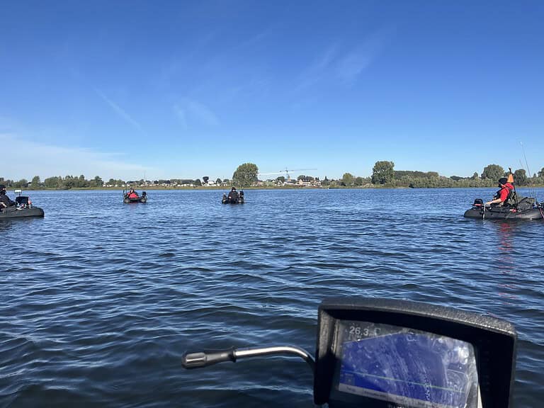 Meerdere mensen in kleine bootjes vissen op een kalm meer onder een helderblauwe lucht met een stad zichtbaar op de achtergrond. Het controleapparaat van een boot is op de voorgrond, ongeveer twee meter verderop.