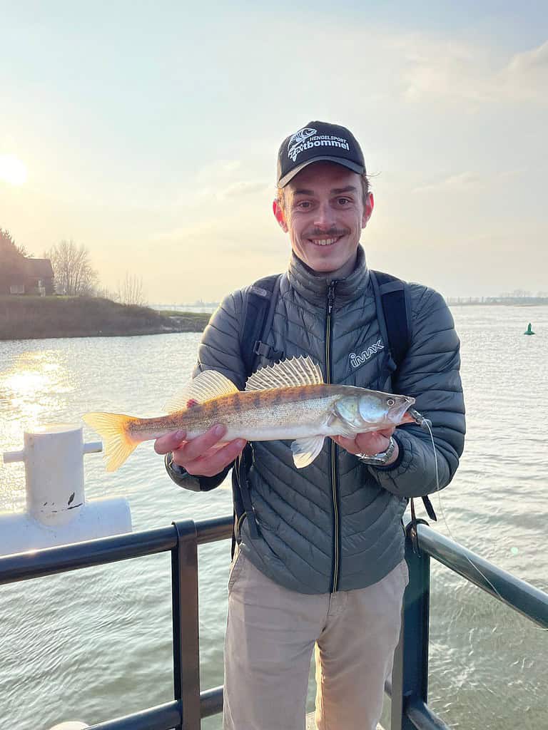 Een persoon met een pet en jas staat op een steiger met een vis in zijn handen, die hij waarschijnlijk heeft gevangen tijdens een middagje Snoekbaarsvissen. Op de achtergrond ziet u de schilderachtige waterkant van de rivier.