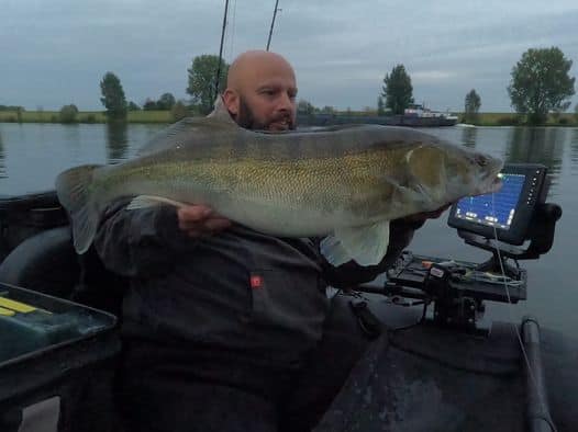 Een persoon zit in een boot met een grote vis in zijn handen. Ernaast is een fishfinder-scherm zichtbaar op een kalm wateroppervlak op de achtergrond.