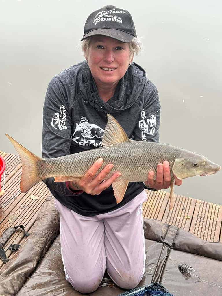Cindy Horstman knielt op de kade, met trots een grote barbeel in haar handen. Ze draagt een pet en een donker shirt met lange mouwen, met een uitgestrekt wateroppervlak achter haar.