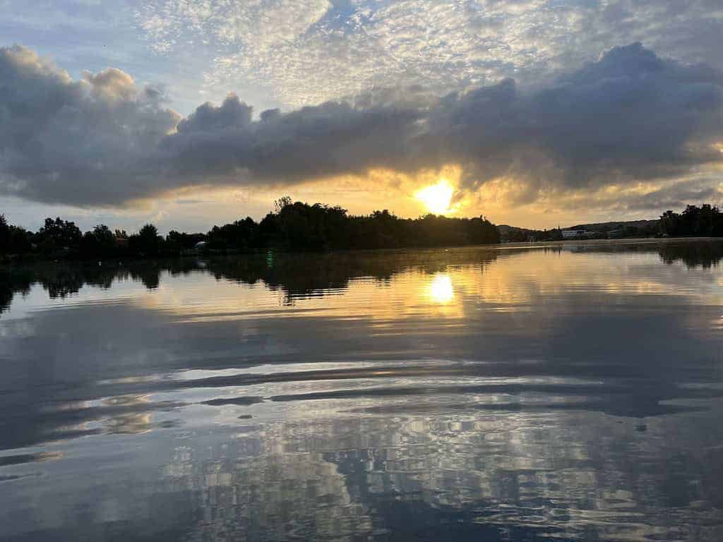 Zonsopgang boven een rustig meer in de Lot-regio in Frankrijk, met silhouetten van bomen aan de horizon en wolken in de lucht die worden weerspiegeld in het water. Dit creëert een serene achtergrond voor iedereen die op zoek is naar een ochtendavontuur waarbij u op zwarte baars vist.