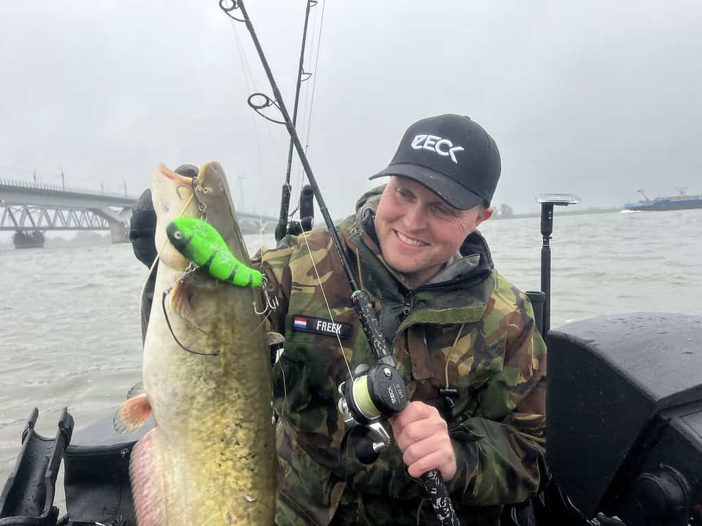 Een persoon op een boot, gekleed in camouflagekleding en een zwarte pet, houdt een grote vis omhoog die hij met een groen lokaas heeft gevangen. De achtergrond toont een watermassa en een brug.