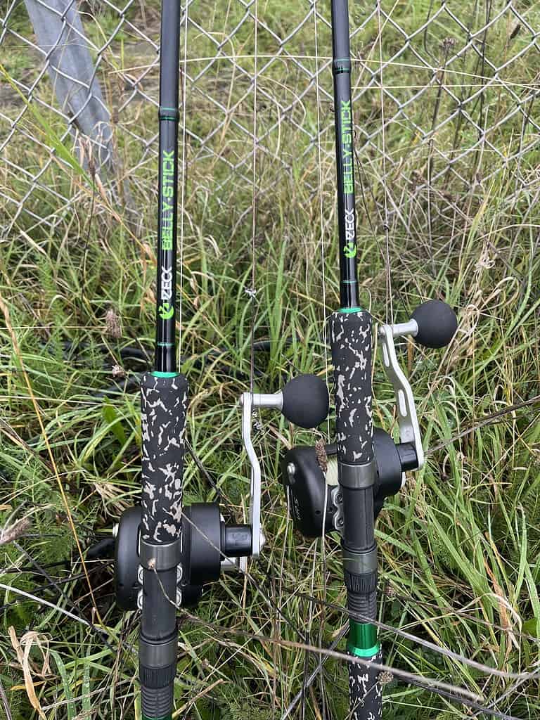 Twee camouflage-gepatroneerde hengels met molens staan op een grasveld bij een hek van gaas. De hengels zijn gelabeld als "Zebco Beast Stick".