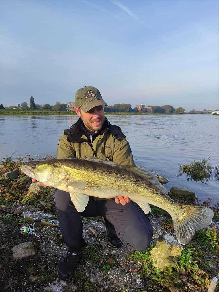Een persoon houdt trots een grote vis vast bij de rivier, met een pet en buitenkleding. De scène legt de essentie van "vangsten van de week" vast, met glinsterend water onder een heldere hemel en verre gebouwen die bijdragen aan de pittoreske achtergrond.