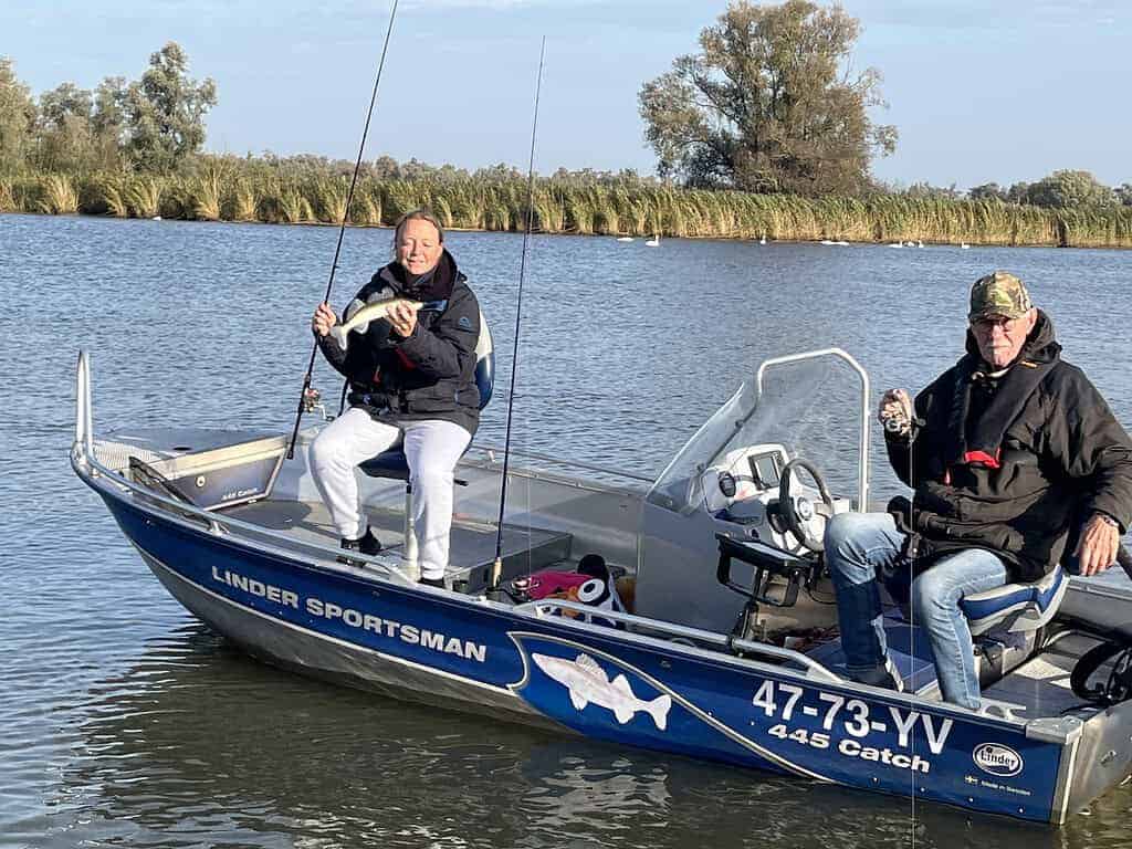 Twee mensen vissen op een blauwe boot met het label "Linder Sportsman 447 Catch" in een kalm waterlichaam, omringd door hoog gras en bomen. Een van hen houdt een hengel vast; beiden dragen jassen.