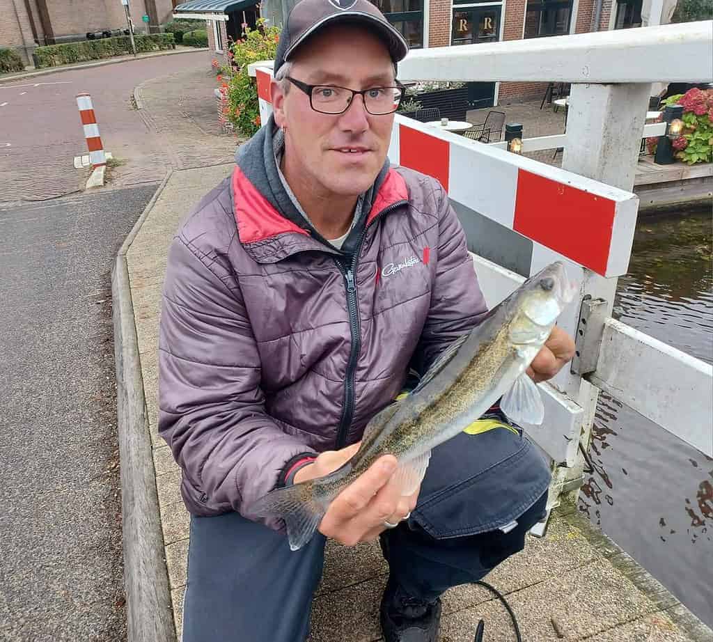 Een man knielt trots op een brug en toont zijn vangsten, met het serene water en de torenhoge gebouwen als achtergrond.