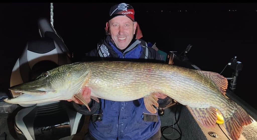 Een persoon staat 's nachts trots op een boot, met in zijn hand de indrukwekkende vangsten van de week - een grote vis - terwijl hij een pet en een jas draagt.