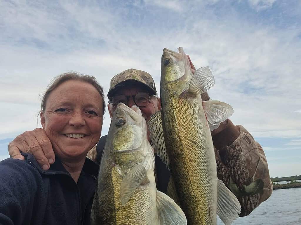 Twee mensen houden trots hun vangsten van de week omhoog – twee grote vissen – en lachen buiten onder een bewolkte lucht.