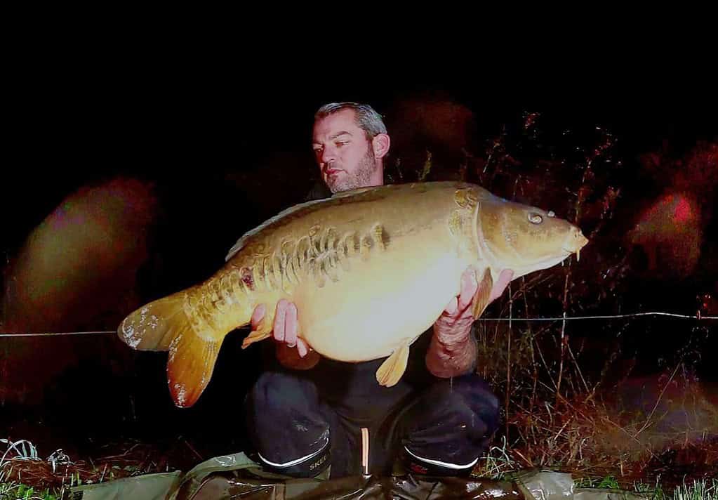 Een persoon toont trots zijn vangsten 's nachts, knielend op een mat, tegen een donkere achtergrond.