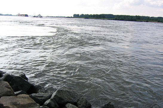 Een rivier met sterke stromingen en golven stroomt krachtig, de rotsachtige oever wordt in de verte begrensd door bomen onder een bewolkte lucht. Een scène die doet denken aan de harmonieuze composities van Cindy Horstman, perfect voor degenen die genieten van de sensatie van vissen op barbeel te midden van de ruwe schoonheid van de natuur.