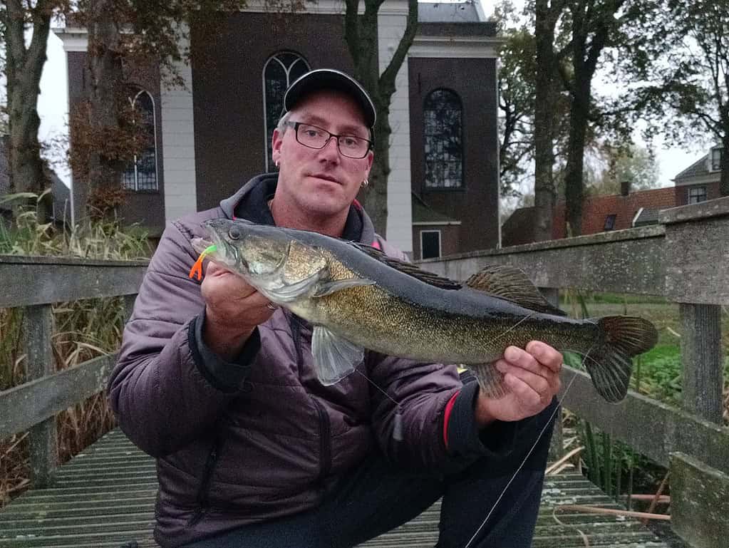 Een persoon die trots een grote vis vasthoudt op een houten brug, tandjes die glinsteren van plezier, terwijl een pittoresk kerkje en weelderige bomen het perfecte decor vormen voor dit opmerkelijke vangsten-moment.
