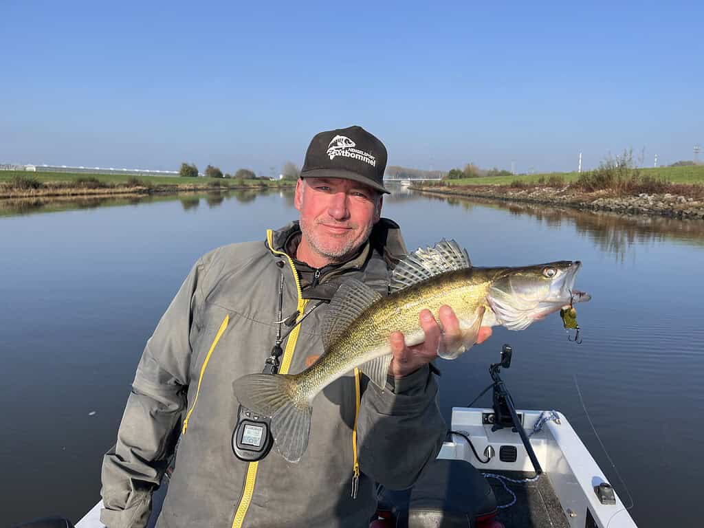 Joris en Johan, gekleed in een grijs jasje en pet, demonstreren vakkundig de kunst van het baarzen vangen terwijl ze een vis vasthouden in hun bootje in een kalme rivier.