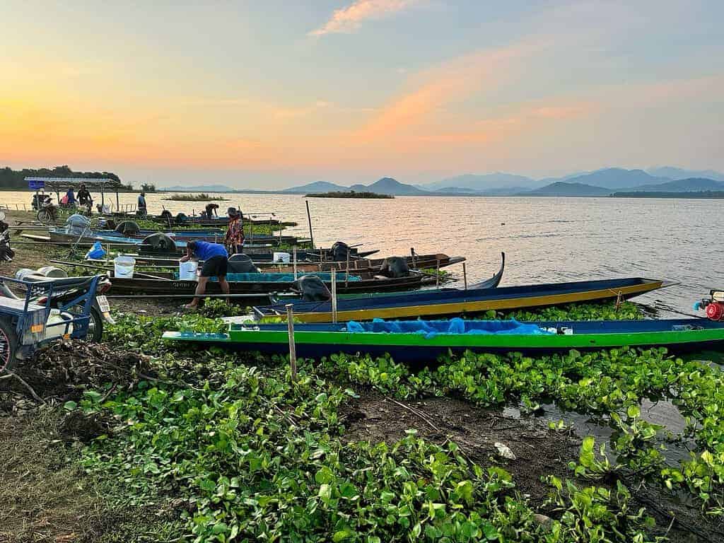 Boten aangemeerd aan een serene oever van een meer in Thailand, met weelderig groen en majestueuze bergen die zich aftekenen tegen de zonsondergang. Mensen, waaronder Mo Simon, verzorgen de boten terwijl het rustige tafereel zich ontvouwt - een perfect einde van een dag waar zelfs een nieuwsgierige slangenkop even bij stil zou kunnen staan.