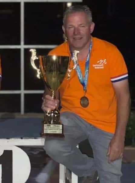 Jan-Willem Plekkenpol, een man in een oranje shirt, draagt trots een grote gouden trofee en draagt een medaille om zijn nek terwijl hij op een podium zit om zijn prestatie als nieuwe eigenaar te vieren.