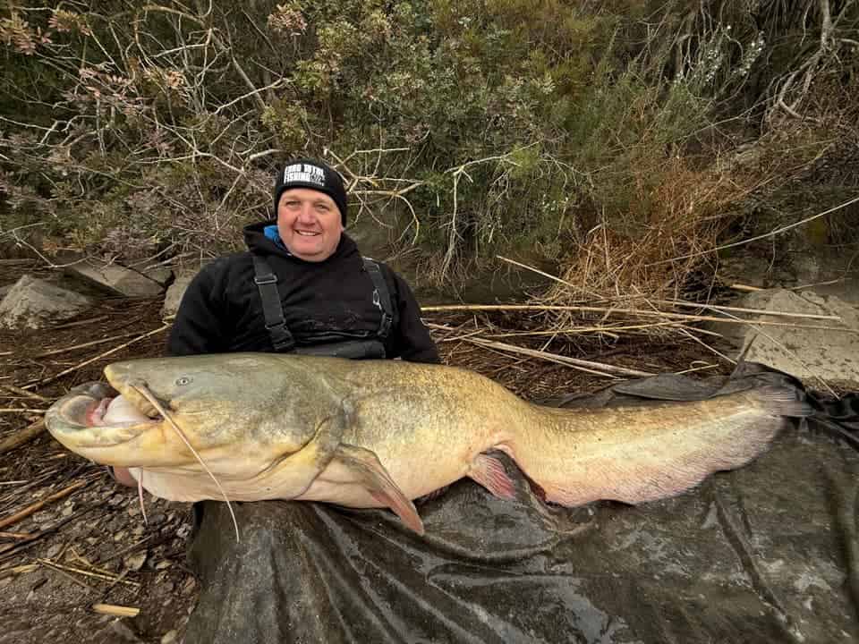 Een persoon toont trots zijn vangsten, een grote meerval, op een zeildoek, terwijl hij op de grond zit met weelderig gebladerte op de achtergrond.