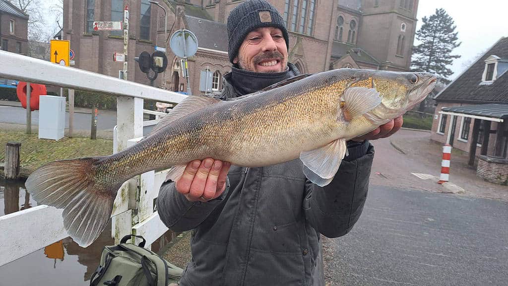 Gehuld in winterkleding toont een persoon trots een grote vangsten tegen de winterse achtergrond van gebouwen en een eenzame boom. Een vleugje Blue Monday hangt in de lucht en geeft diepte aan dit serene tafereel.