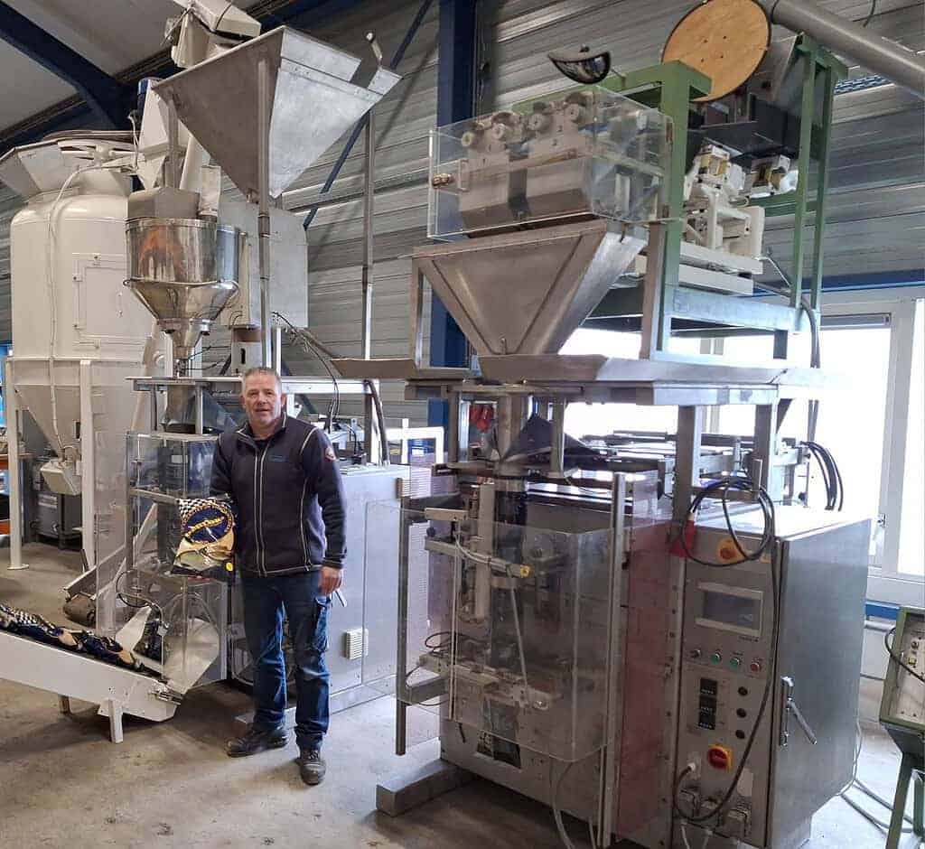 Jan-Willem Plekkenpol staat naast geavanceerde industriële verpakkingsmachines in een drukke fabrieksomgeving en houdt met veel zelfvertrouwen en oog voor detail toezicht op de operatie.