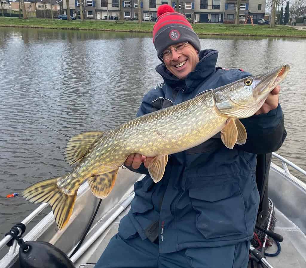 Een persoon in een boot houdt een grote vis vast en lacht naar zijn vangsten van de week. Ze dragen een bril, een rood-grijze muts en een blauw jasje. Een watermassa en gebouwen vormen de achtergrond van dit levendige tafereel.