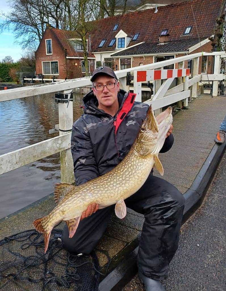 Een man knielt op een pad langs het kanaal en toont trots zijn vangsten (de vangst van de week), een grote vis, met op de achtergrond een charmant huis en rustig water.