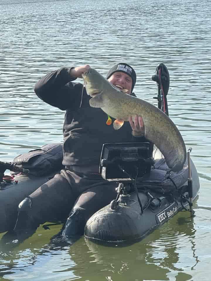 In een wetsuit zit een persoon trots in een opblaasboot en laat zijn indrukwekkende roofvisvangst van de week zien: een enorme meerval die uit het glinsterende water is gevist.