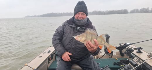 Een persoon in een donker jasje en een muts houdt trots een roofvis vast op de boot, terwijl de bewolkte lucht en het water het perfecte decor vormen voor volle bak vangsten.