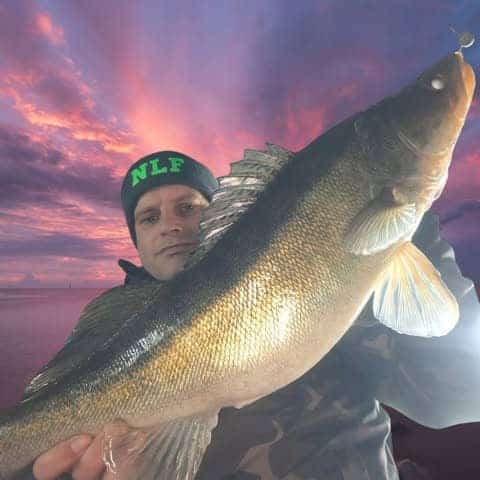 Een persoon toont trots een grote roofvis tegen een stralende zonsondergang en viert met vreugde dat hij zijn vangsten heeft behaald.