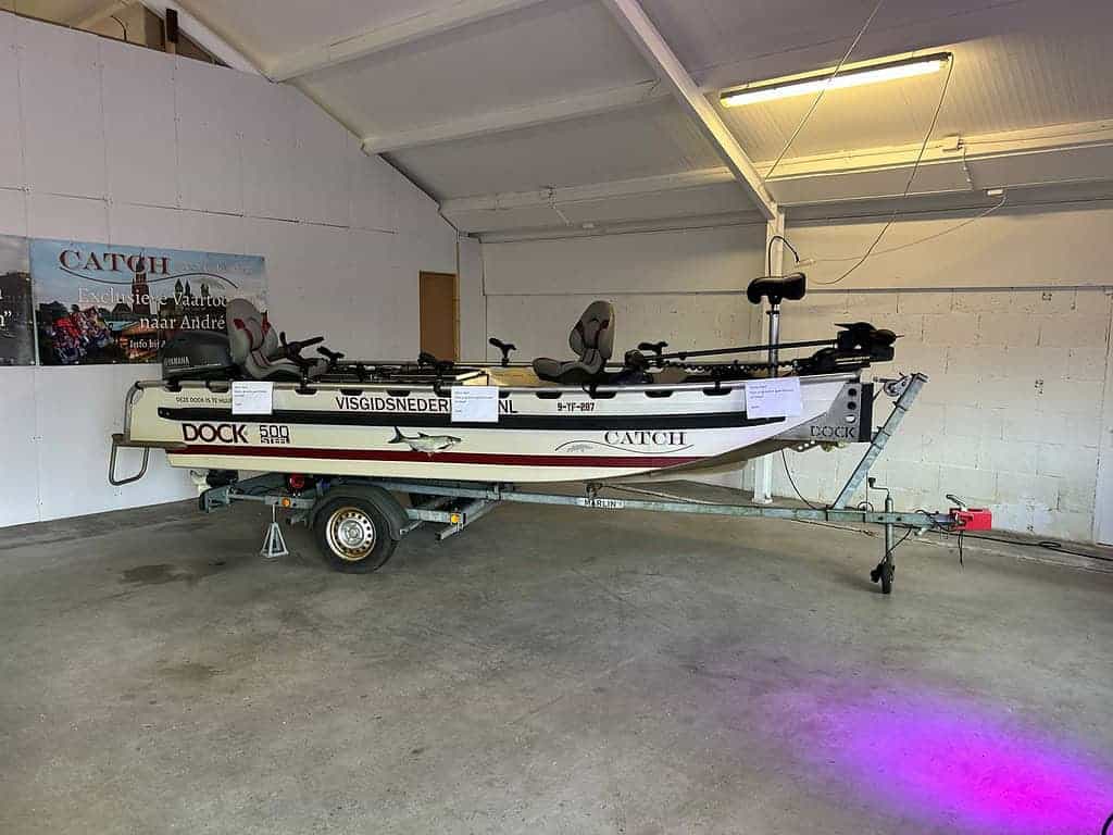 In een garage rust een boot op een trailer, versierd met visgerei en reclameborden voor Hengelsport Maarssen. De scène vangt de essentie van Gezelligheid, ter voorbereiding op een spannende Open Roofvis Dag op het water.