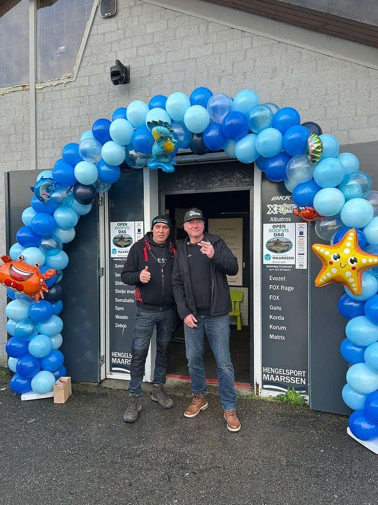 Twee mensen staan voor een deuropening versierd met blauwe en witte ballonnen en opblaasbare zee-thema's, die de gezelligheid van Open Roofvisdag belichamen. Beiden geven een duimpje omhoog, waarmee ze de geest van Hengelsport Maarssen vastleggen.
