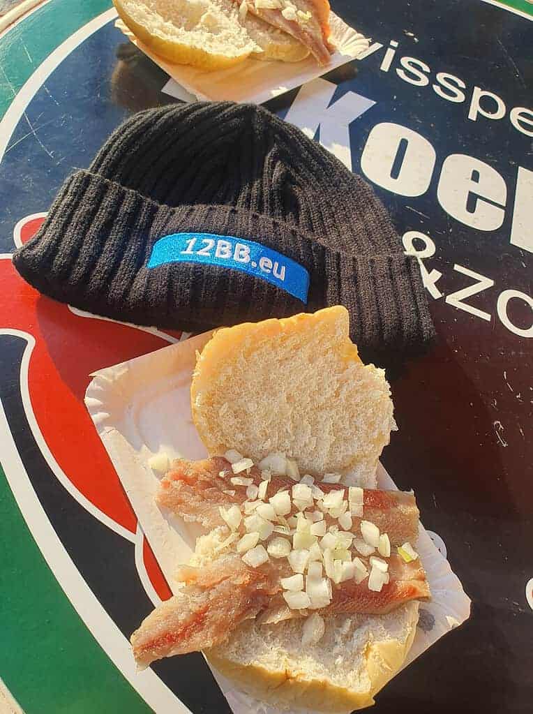 Een broodje met rauwe vis en gesneden uien, een waar genot, ligt op een papieren bordje naast een zwarte muts met daarop "12BB.eu", het geheel is kunstig uitgestald op een kleurrijke ronde tafel.