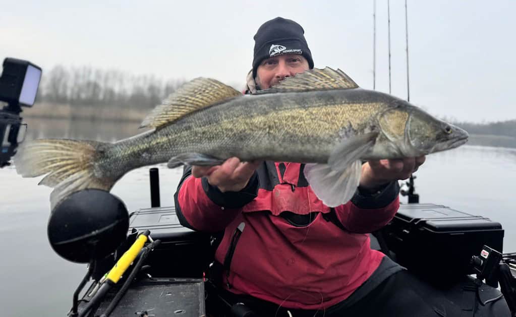 Een persoon in een rood jasje toont triomfantelijk zijn vangsten, met een grote vis in de boot. Visgerei omringt hen terwijl ze te midden van het serene mistige meer op de achtergrond staan.