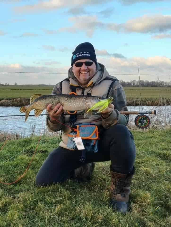 Knielend op het gras bij een serene rivier, draagt de persoon een muts, zonnebril en een jas terwijl hij trots zijn "visje" van de week vasthoudt. Achter hen ligt hun vertrouwde hengel, wat een andere succesvolle entree markeert tussen hun vangsten.