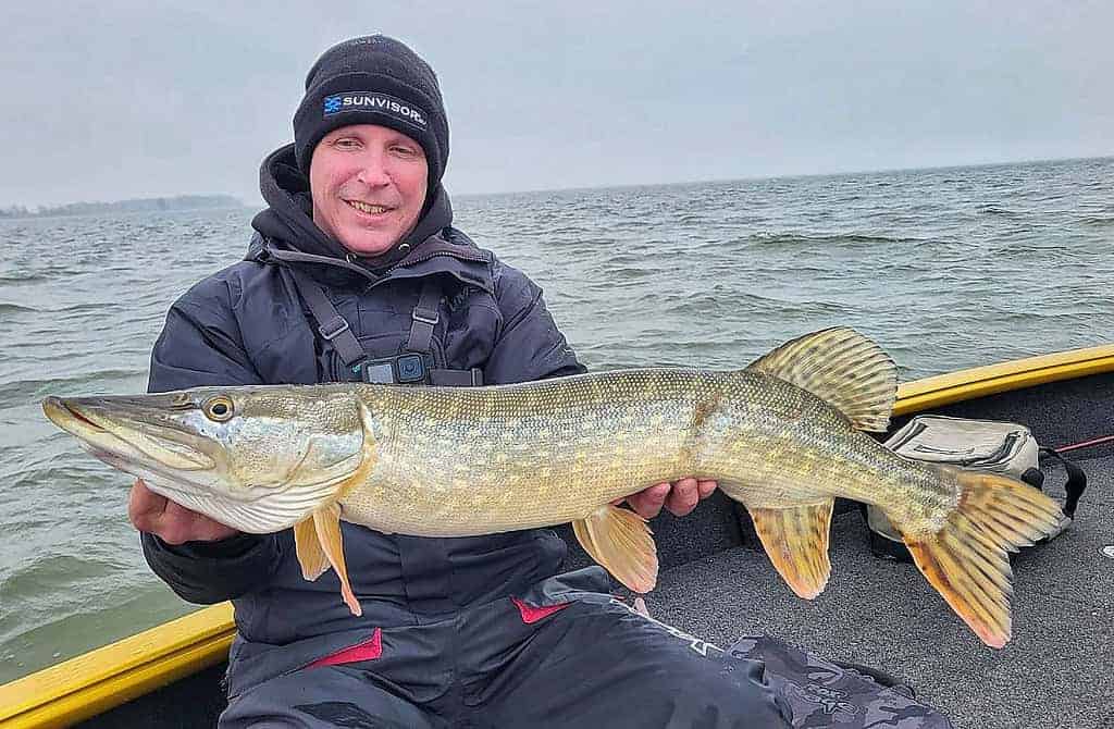 Persoon op een boot die een grote vis vasthoudt tegen een achtergrond van een bewolkte lucht en water, die trots de vangsten laat zien. Gekleed in Predatorgear's zwarte jas en hoed, belichamen ze de sensatie van de vangst.