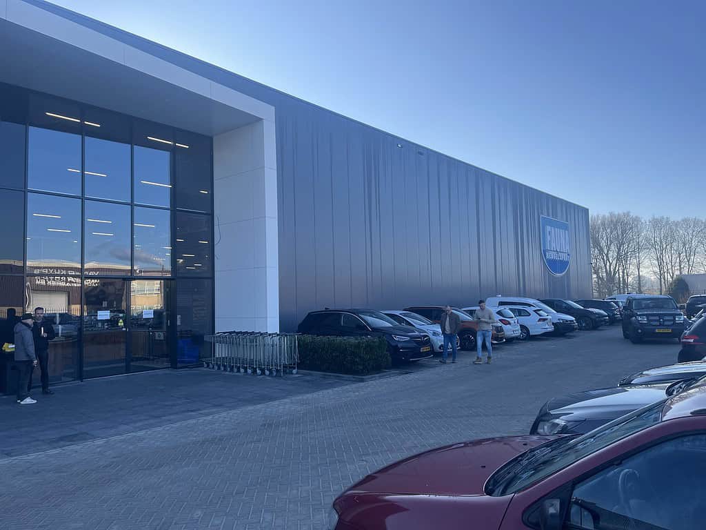 Mensen lopen naast de grote moderne megastore met geparkeerde auto's ervoor. Winkelwagentjes staan in de rij bij de ingang, terwijl Fauna 3.0 zich voorbereidt om zijn deuren te openen. Heldere lucht boven ons hoofd.