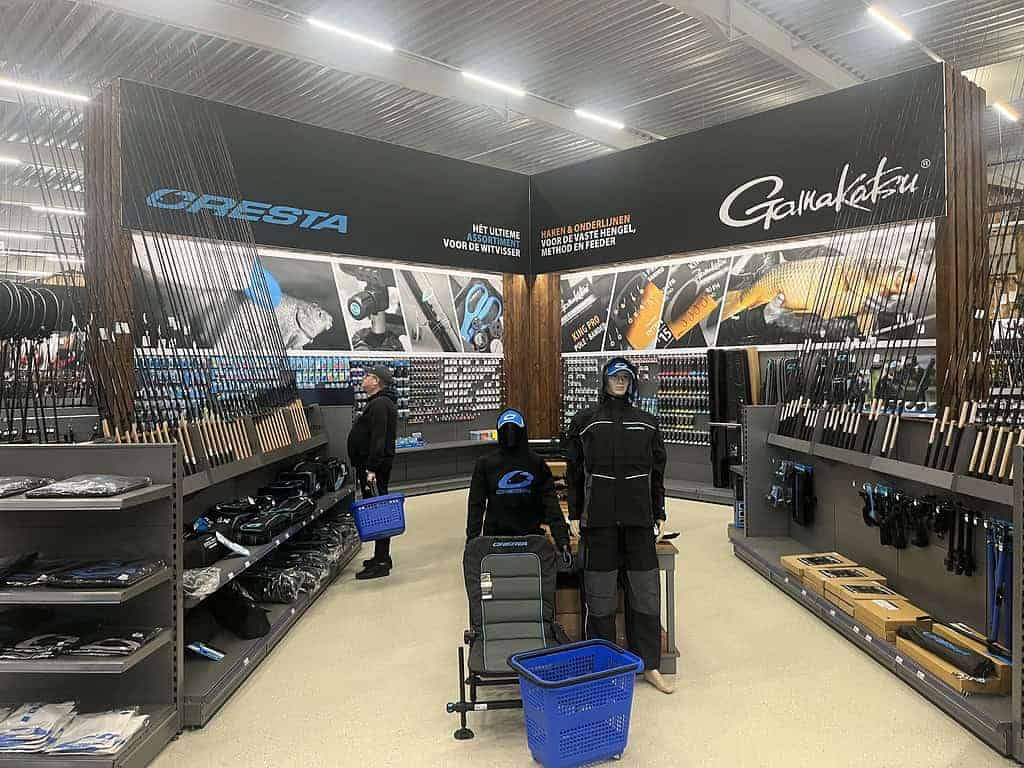De nieuwe megastore heeft een gangpad met visgerei met de nieuwste Fauna 3.0-hengels, stijlvolle kleding en essentiële accessoires met levensechte paspoppen. Klanten met blauwe manden bekijken aandachtig de selectie onder felle plafondlampen.