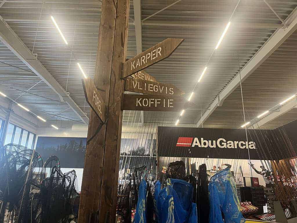 Houten bordjes op een paal in de megastore verwijzen naar verschillende afdelingen, waaronder Karper, Zeebaars, Vliegvis en Koffie. Een display met vistuig en een Abu Garcia-sectie zijn zichtbaar terwijl Fauna 3.0 overal zijn deuren opent voor liefhebbers.