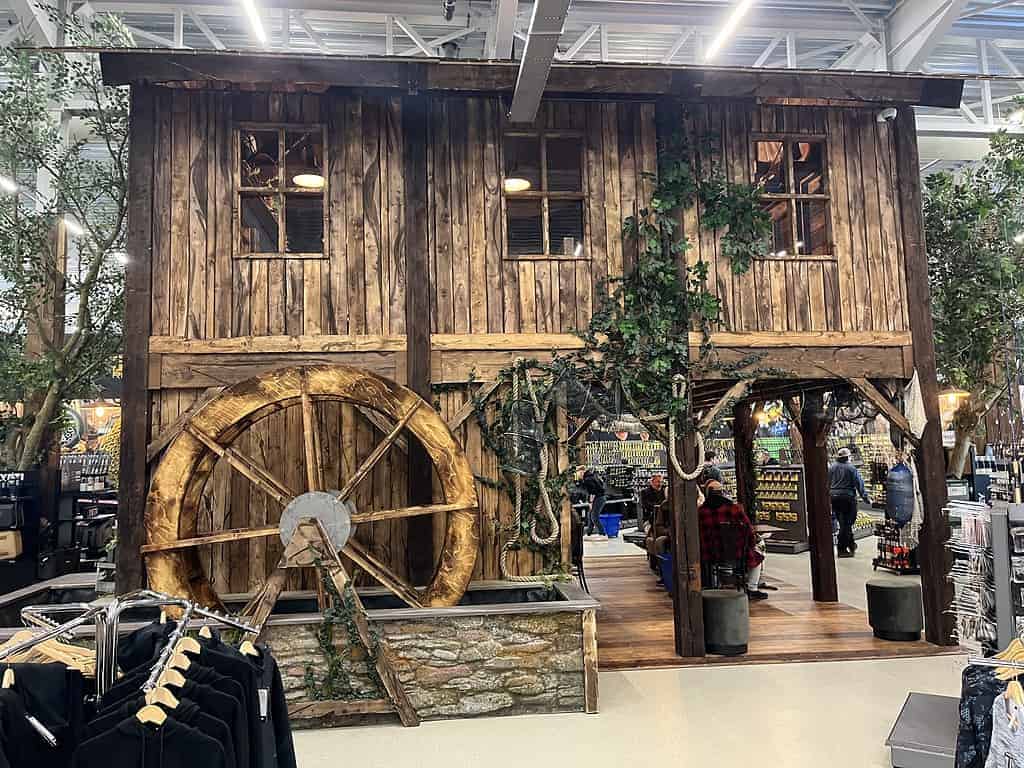 Binnen staat een houten watermolen met een groot wiel, verscholen tussen kunstmatig groen en kledingrekken. Het doet denken aan de Fauna 3.0-ontwerpstijl die je zou verwachten in een megastore die klaar is om zijn deuren te openen.