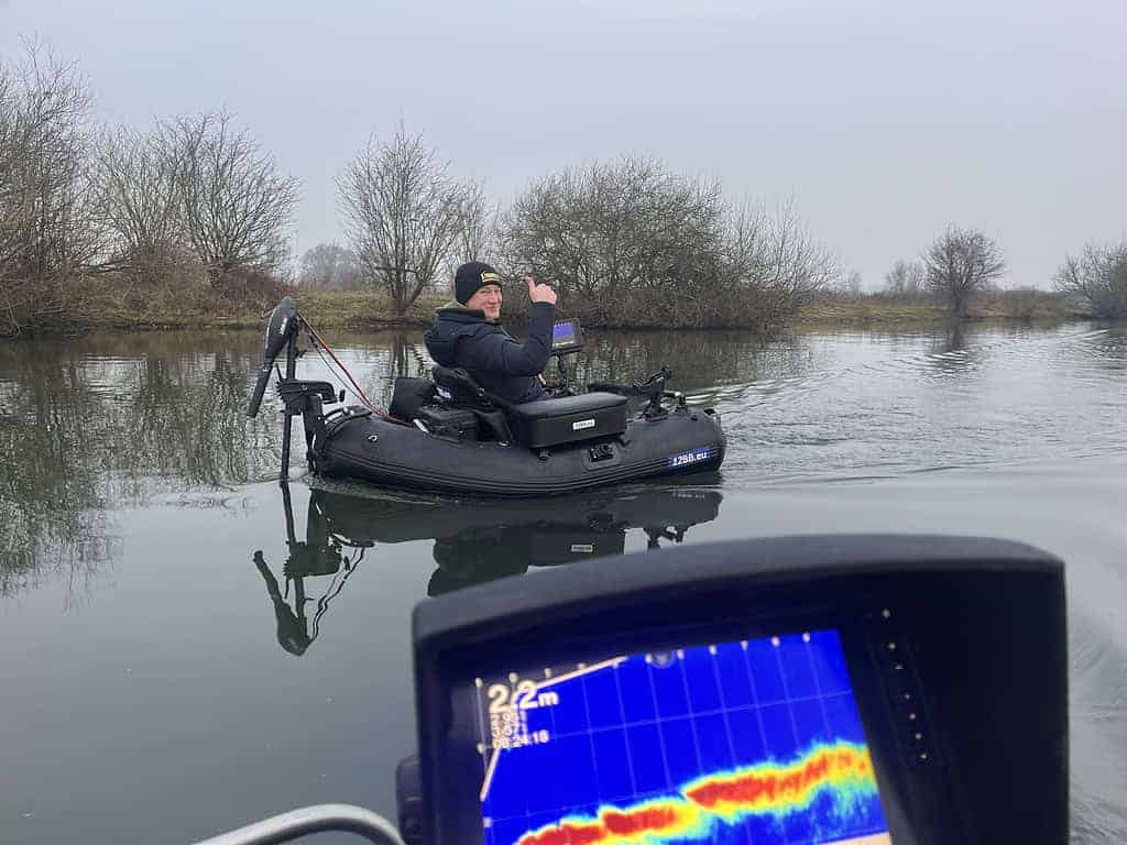 Een persoon in een klein bootje op een kalme rivier gebruikt een apparaat om de activiteit van vissen in de gaten te houden. Op de voorgrond worden onderwatergegevens weergegeven op het scherm. Het serene tafereel wordt vastgelegd alsof het onderdeel is van een exclusieve video die gratis wordt weggegeven.