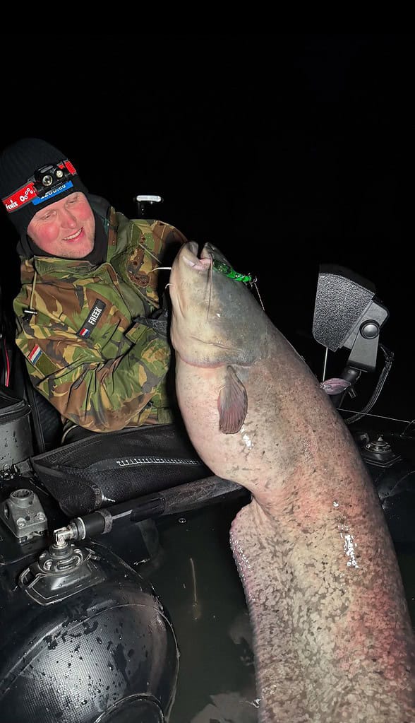 Persoon in camouflagejas en hoofdlamp die 's nachts op een boot een grote vis vasthoudt.